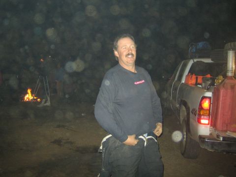1990 Ford Bronco - grinning man in a dark pullover stands beside a pickup truck at a nighttime desert camp, a campfire glowing behind him.