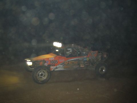 1990 Ford Bronco - orange off-road race buggy marked 1005 speeds through the dark with headlights blazing during a night desert race.