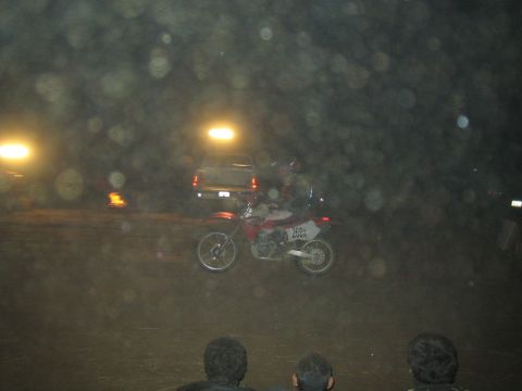 1990 Ford Bronco - red dirt bike races across a dark dirt track at night as silhouetted spectators and a parked SUV look on.