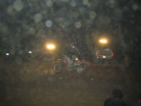 1990 Ford Bronco - helmeted rider pilots dirt bike E11 across a dim dirt course at night, headlights and a parked SUV glowing behind.