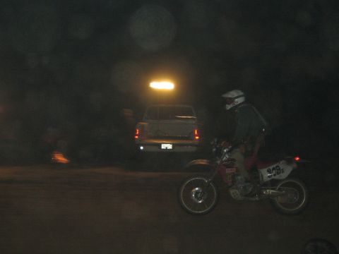 1990 Ford Bronco - helmeted rider on dirt bike 326 idles in the dark next to a light-colored pickup truck at a night desert event.