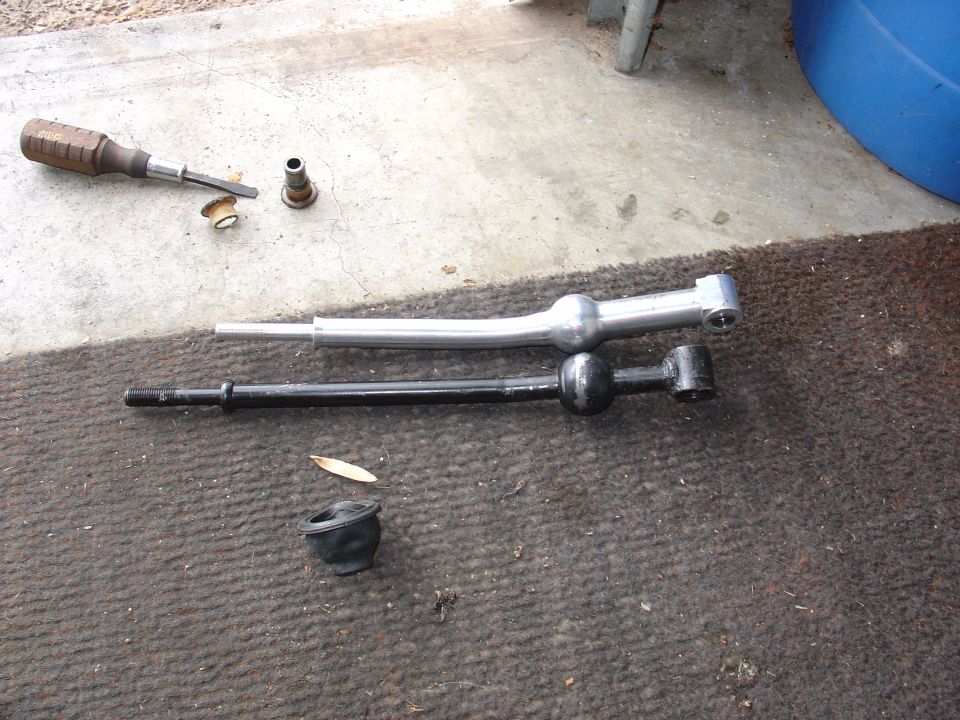 Polished aluminum short shifter next to black stock shifter lever on carpet, with screwdriver and rubber boot nearby