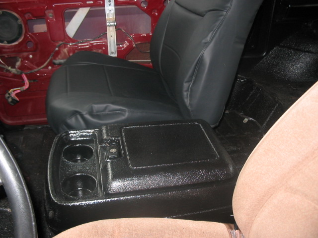 Interior shot of a vehicle showing a black vinyl driver's seat, a black center console with cup holders, and the red door panel with exposed wiring