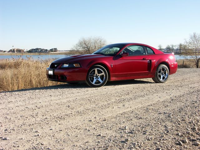 Red coupe parked on a gravel road near a body of water
