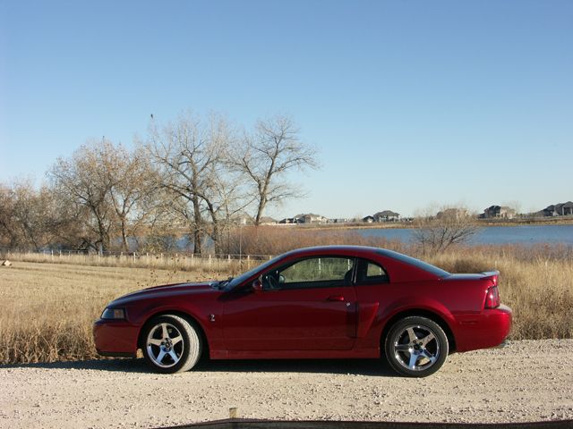 Red coupe parked on a gravel road with a lake and houses in the background