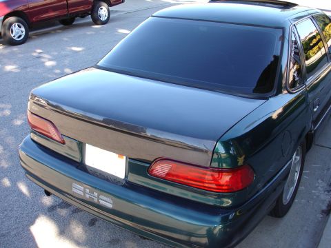 1993 Ford Taurus - The rear of a dark green sedan is shown, a carbon fiber trunk lid accent and tinted windows. A red pickup truck is visible in the backgroun…