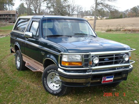 1995 Ford Bronco - A dark green and tan Ford Bronco is parked on a grassy area with trees in the background.
