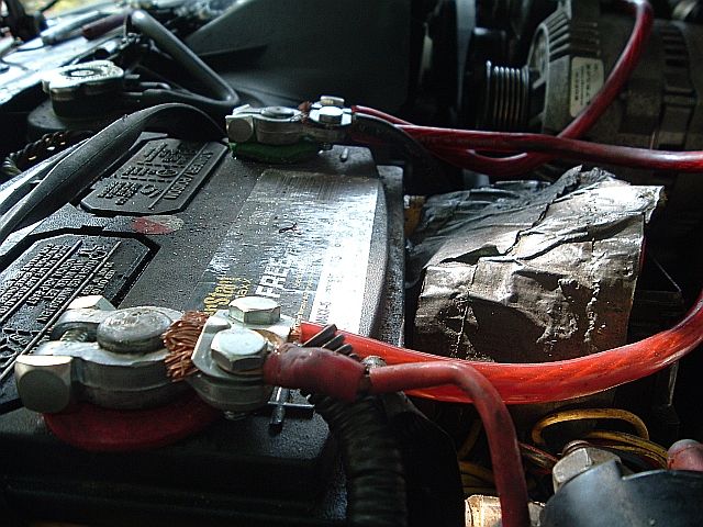 Battery top with both terminals clamped, red cables, and a foil-wrapped component behind