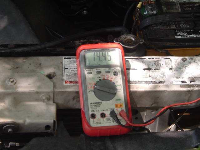 Red Craftsman multimeter reading 14.45 volts resting on a radiator support in an engine bay