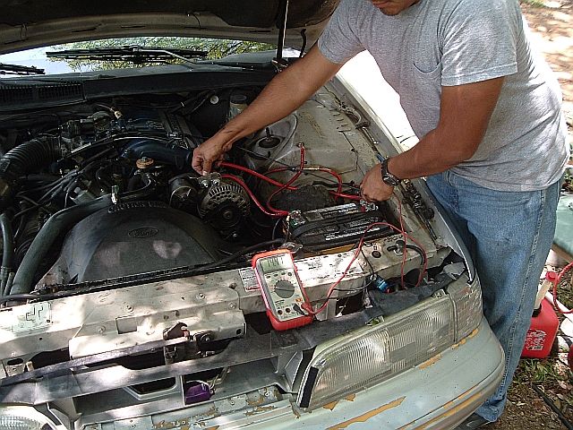 Man in gray shirt leaning over a Ford engine bay testing the battery with a multimeter