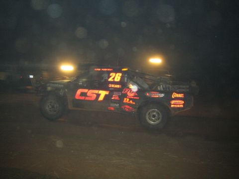 1990 Ford Bronco - black off-road race truck numbered 26 with CST and sponsor decals races through dust under night lights.