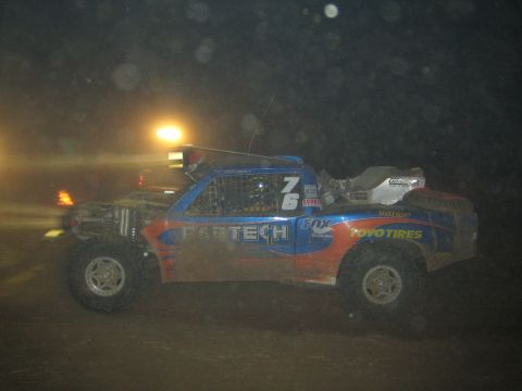 1990 Ford Bronco - blue and red Fabtech-liveried race truck numbered 76 powers across a dark course at a night desert race.