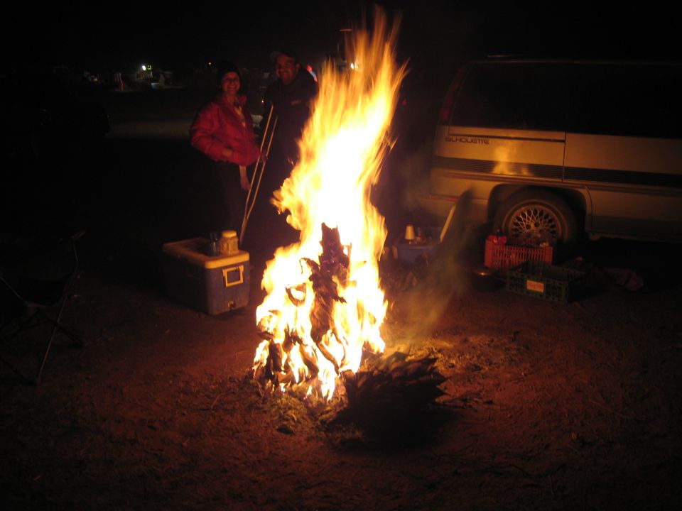 Tall campfire burning at night beside a light-colored van with two people standing nearby