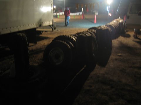 1990 Ford Bronco - Rows of off-road truck tires are staged at night beside a white box trailer, with cones and a person in the background.