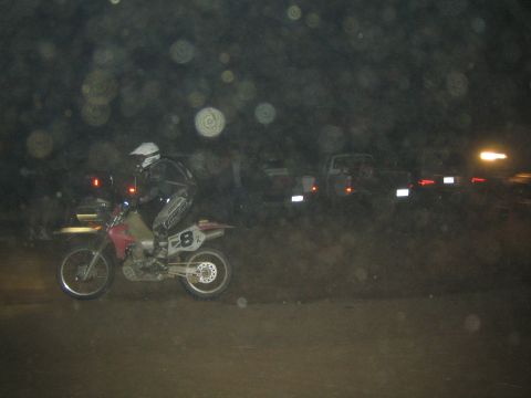 1990 Ford Bronco - helmeted rider on dirt bike 8 races a dark course at night with parked vehicles and taillights glowing behind.