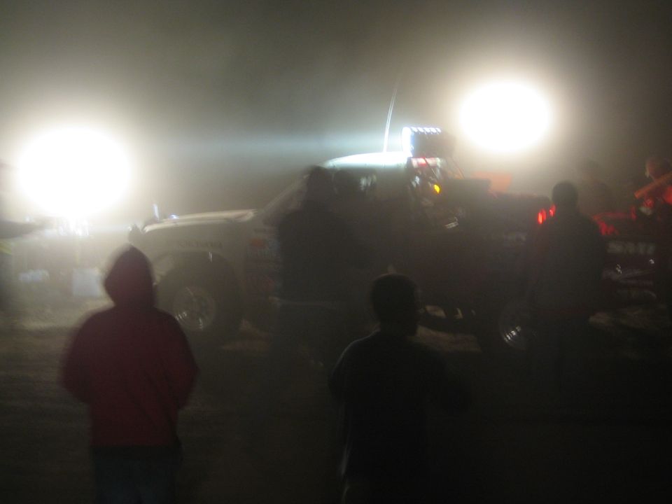 Silhouetted spectators watching a brightly lit off-road vehicle in dust at night