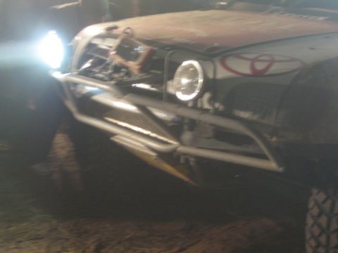 1990 Ford Bronco - Close-up of a race truck's tubular front bumper and round headlight, with a Toyota emblem visible on the grille at night.