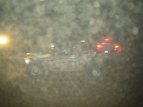 1990 Ford Bronco - off-road race buggy speeds through thick dust at night, headlights glowing as red taillights trail behind.
