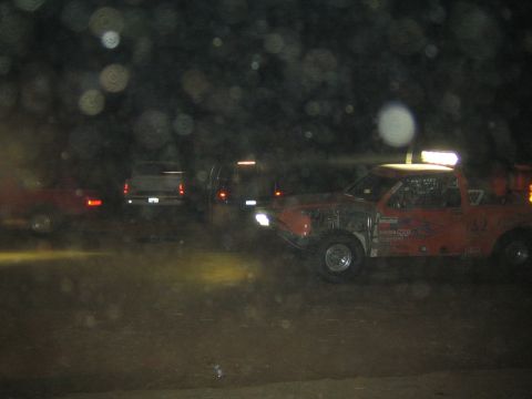 1990 Ford Bronco - orange off-road race truck with a lit roof light bar sits in the dark beside parked trucks at a night desert event.
