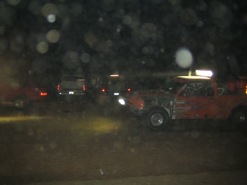 Orange off-road race truck with roof light bar at night beside other parked vehicles