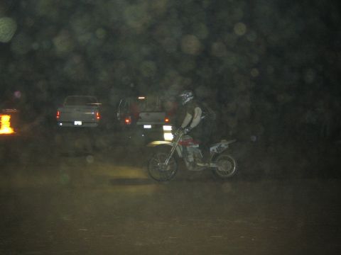 1990 Ford Bronco - helmeted rider on a yellow dirt bike pauses on a dark course at night with two pickup trucks parked behind.
