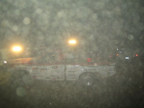 1990 Ford Bronco - white sponsor-covered off-road race truck sits in profile at night, lit by glowing headlights through a dusty haze.