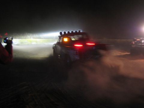 1990 Ford Bronco - off-road race truck with a roof light bar and glowing taillights throws up a cloud of dust on a dark desert course.