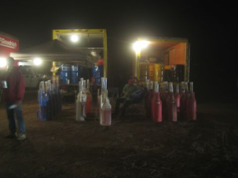 1990 Ford Bronco - Rows of red and blue liquid-filled bottles line a lit pit area at a desert race at night, with a person standing at left.