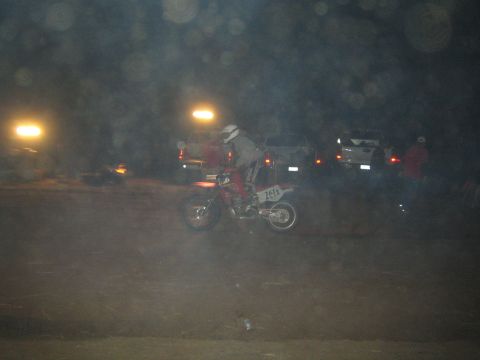 1990 Ford Bronco - motorcyclist on a number 76A dirt bike rides through a dusty night course past parked vehicles and standing spectators.