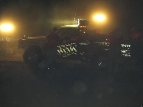 1990 Ford Bronco - off-road race truck bearing a Milgard Windows decal sits in a pit lit by bright floodlights at night, with crew nearby.