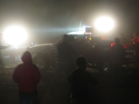 1990 Ford Bronco - Silhouetted spectators in hooded jackets watch a brightly floodlit off-road race truck with a light bar at night.