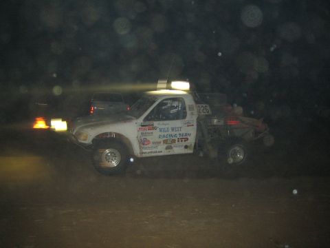 1990 Ford Bronco - white off-road race truck marked Wild West Racing Team and number 225 sits in profile at night, its headlights glowing.