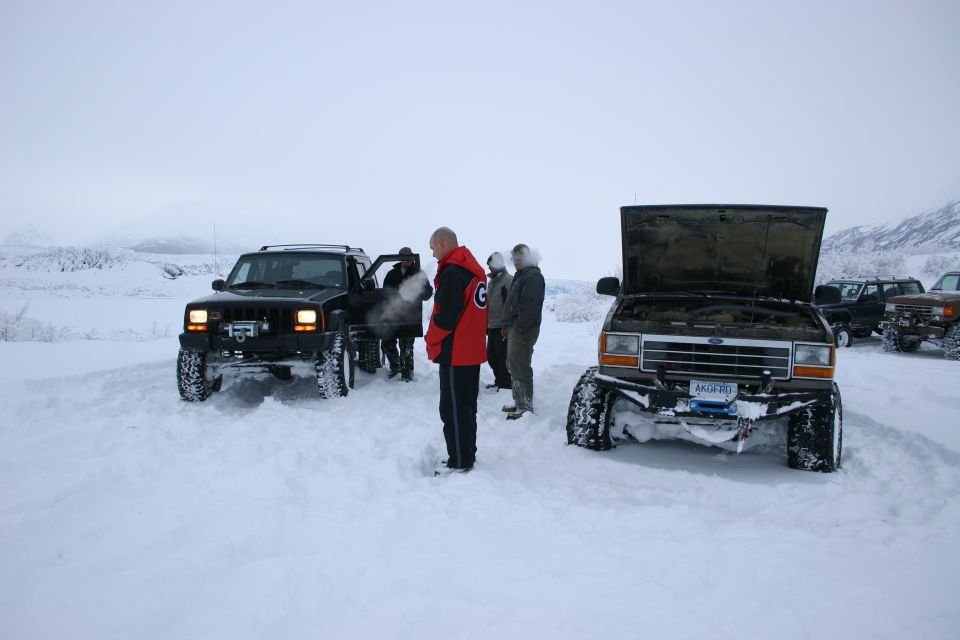 Several people stand around off-road vehicles stuck in the snow. One vehicle has its hood up
