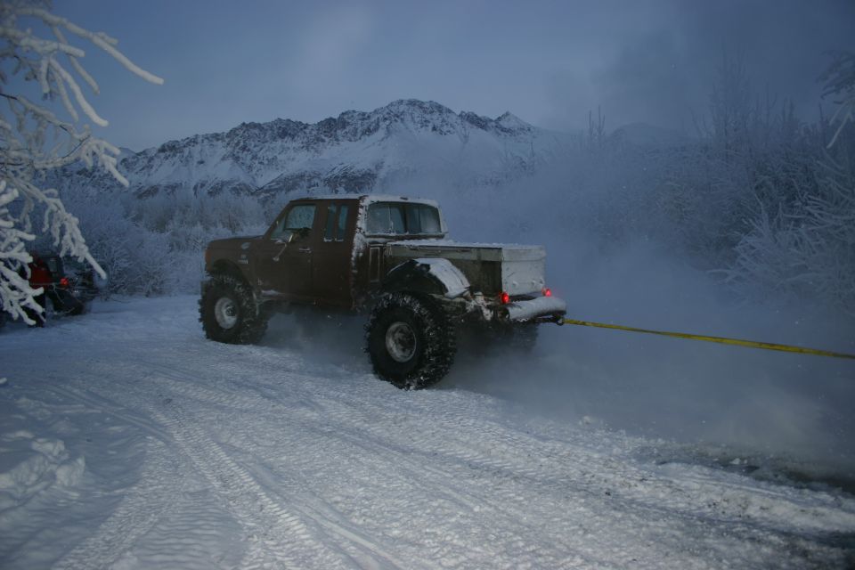 Brown pickup truck with large tires, towing a yellow strap in a snowy landscape with mountains