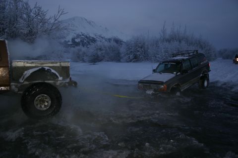 2003 Jeep TJ - A dark SUV is being towed through icy water, with snow-covered trees and mountains visible in the background.