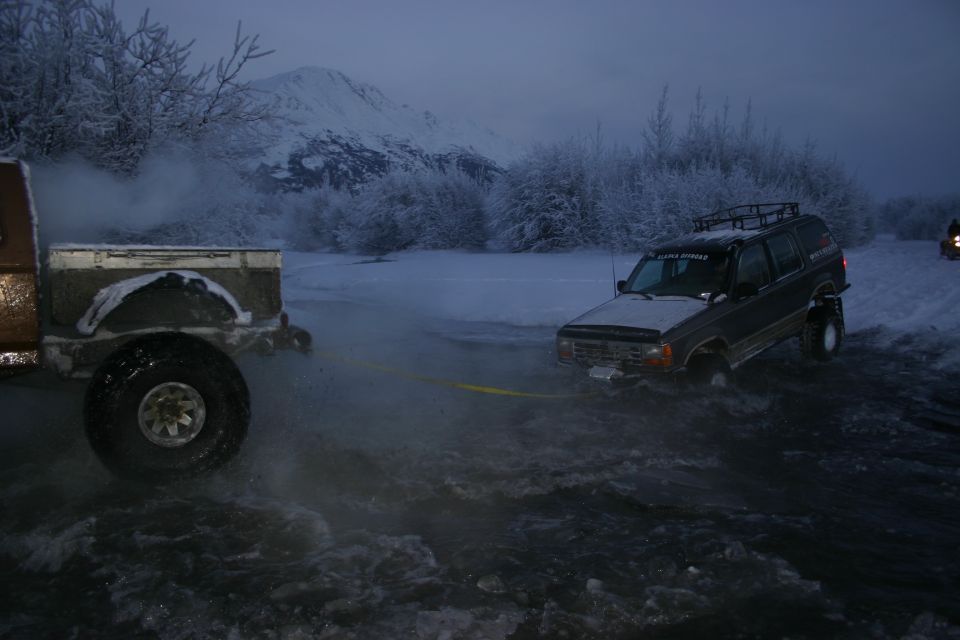 Dark SUV being towed through water, snow-covered trees and mountains in background