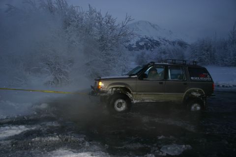 2003 Jeep TJ - Ford Explorer with "SIX STATES" sticker stuck in icy water near snow-covered trees and mountains.