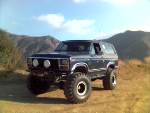 1985 Ford Bronco - Modified Ford Bronco with large tires and off-road lights sits on a dusty trail.