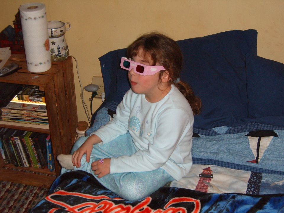 Girl wearing pink 3D glasses sits on a bed, looking to the side. A shelf with books and a roll of paper towels is to her left