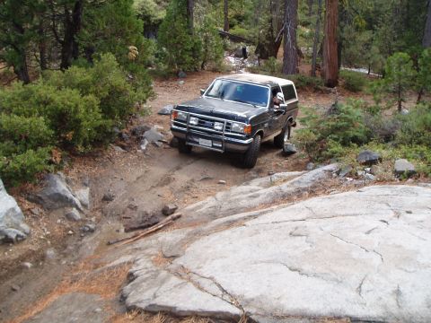 1990 Ford Bronco - This black Ford Bronco navigates a challenging rocky path in a wooded area. Its body style indicates it is likely from the 1987-1991 model…