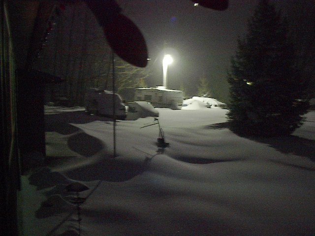 snowy yard at night with a trailer and two covered vehicles under a street light