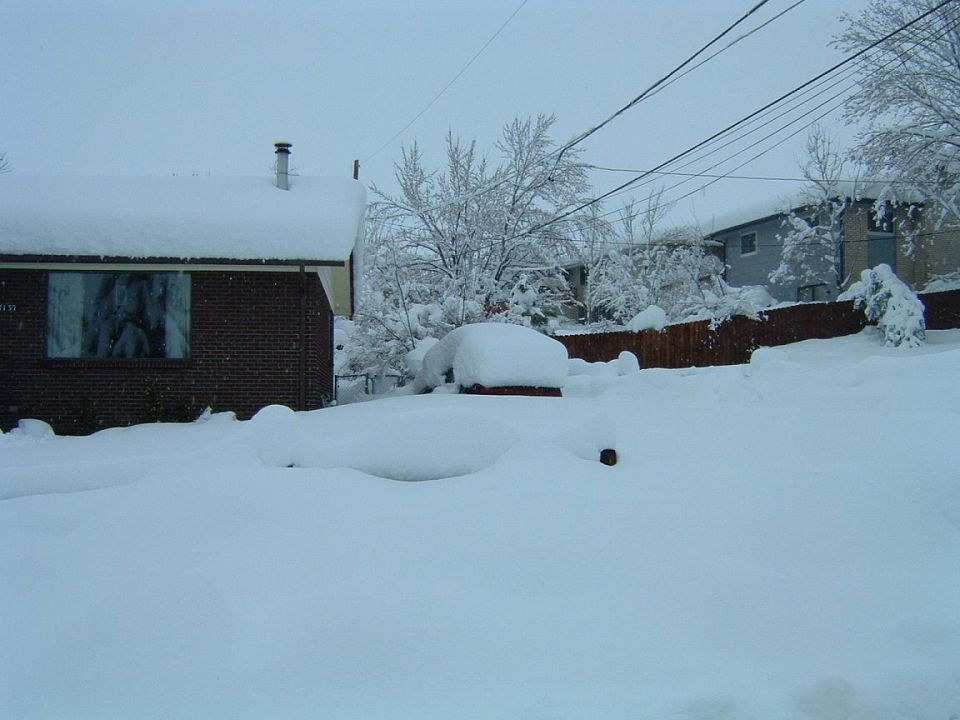Car buried under deep snow next to a brick house, only a sliver of red paint visible