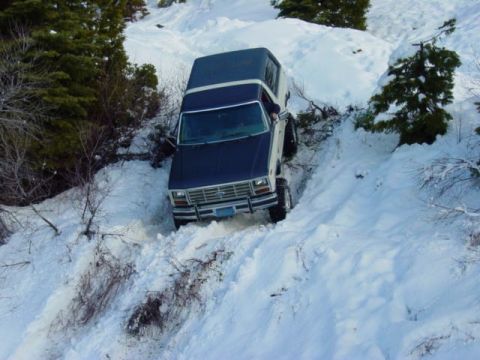 1986 Ford Bronco - A 1986 Ford Bronco tackles a challenging descent down a snow-covered hill, surrounded by trees and wintery terrain.