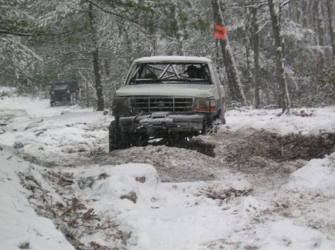 1995 Ford Bronco - A white 1995 Ford Bronco with an external roll cage tackles a muddy, snow-covered trail, with another vehicle visible in the background.