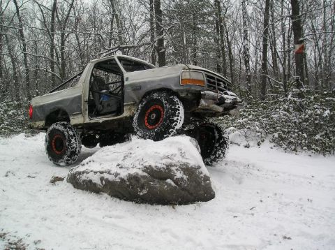 1995 Ford Bronco - A heavily modified truck navigates a snowy obstacle in a wooded area.