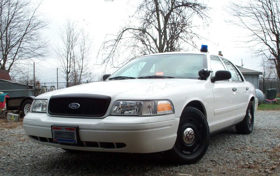 White Ford Crown Victoria police sedan with roof light bar on gravel lot