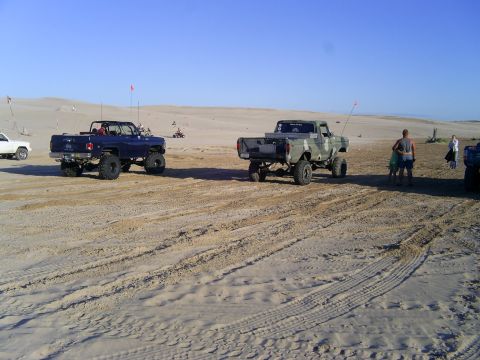 1977 Ford F-150 - Two modified pickup trucks are parked on a sandy terrain, with people and other vehicles in the distance.