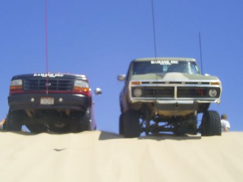 1977 Ford F-150 - Two pickup trucks, one from the 1970s and a more modern one, are positioned on sand dunes under a clear blue sky.