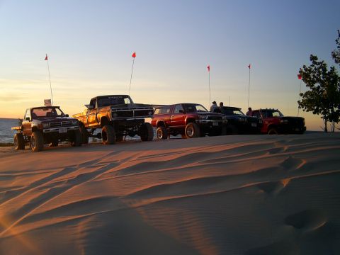 1977 Ford F-150 - Several lifted trucks are parked on sand dunes with red flags flying.
