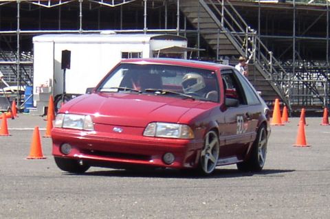 1987 Volkswagen Golf - Red 1987 Ford Mustang hatchback navigates a course marked by orange cones, driver in helmet visible.
